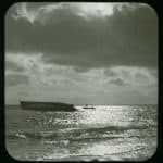 A view from the beach of the hull of a larger boat upturned in the water. To the right of the hull is a small boat with two people aboard. The plate has a sticker top right 'J. Dore, Sandown, IOW'.