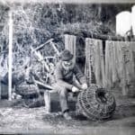 Fishermen sitting making crab pots. Bathing huts can be seen on the hillside above the fishermen