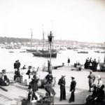 View from Cowes pontoon looking out over the pontoon and water with East Cowes in the background. Numerous people are standing and seated including sailors