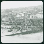 View of Ventnor and its beach viewed from the pier. Queen's Hotel and Esplanade Hotel can be seen, there are large crowds and a number of flags are flying on the seafront