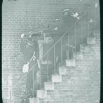 Three ambulancemen carrying a patient down a flight of stairs on a stretcher. Sandown in 1914