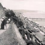 View of Sandown Bay from the cliff path at Sandown. People are walking down the path and one man is pointing at a large crowd gathered below on the Esplanade