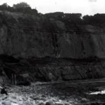 Section of cliff with a steps going up one side. People are on the rocky beach near the steps