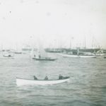 Boats on the water at Cowes. A small launch is in the foreground, whilst a number of large yachts decked with flags are in the background