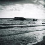 Three men in rowing boat inspecting the upturned hull of wrecked ship in Sandown Bay